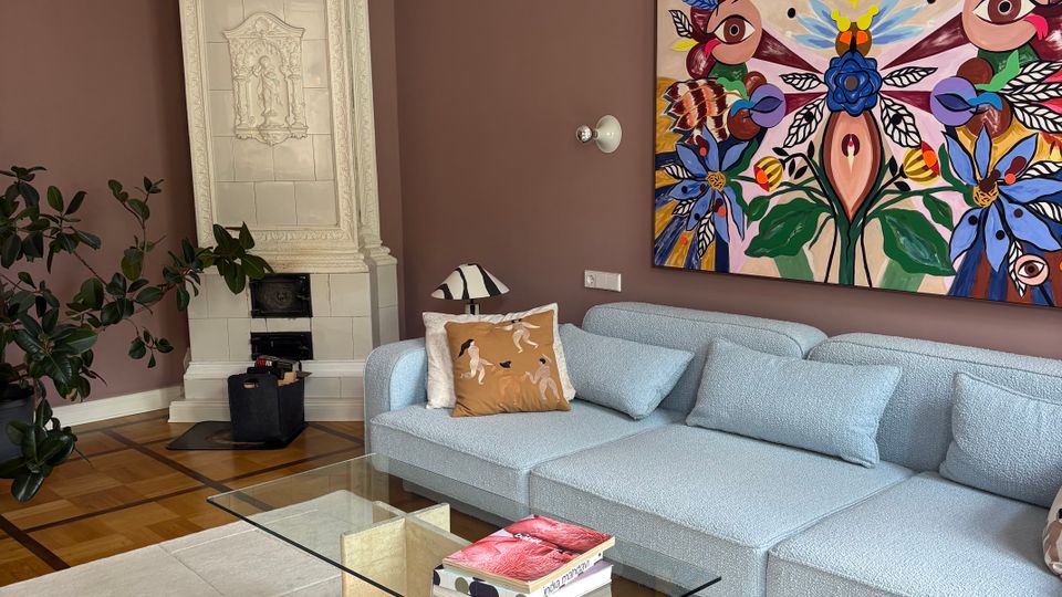 Elegantes Wohnzimmer mit hohen, mauvefarbenen Wänden, Stuckfries und einem dekorativen weißen Kachelofen in der Ecke. Vor einem großen, farbenfrohen Wandbild stehen ein hellblaues Sofa, ein gläserner Couchtisch auf hellem Teppich sowie eine große Hängeleuchte und Parkettboden.