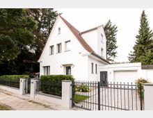 Weißes Einfamilienhaus mit Satteldach und modernen Fenstern, umgeben von einem gepflegten Garten und einer niedrigen Hecke. Ein schmiedeeiserner Zaun und ein gepflasterter Weg führen zu einer schwarzen Haustür und einer angebauten Garage.