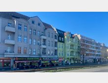 Straßenansicht mit einer Reihe mehrstöckiger Wohnhäuser im Altbaustil, verputzt in Grau und Grün, mit kleinen Balkonen. Im Erdgeschoss befinden sich Geschäfte wie ein Gemüseladen, eine Dönerbude und andere Läden; davor eine breite Straße mit Straßenlaternen.