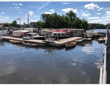 Ein kleiner Hafen mit mehreren Anlegestellen, an denen Boote, Ruderboote und Hausboote festgemacht sind. Im Hintergrund sind Bäume, ein Fluss und zwei Fabrikschornsteine zu sehen, unter einem sonnigen Himmel mit einigen Wolken.