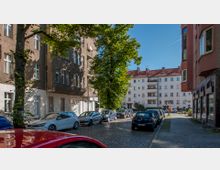 Straßenszene in einer Wohngegend mit mehrstöckigen Wohngebäuden aus Putz und Ziegeln, parkenden Autos auf beiden Seiten der gepflasterten Straße, einem Baum in der Mitte und klarem Himmel im Hintergrund.