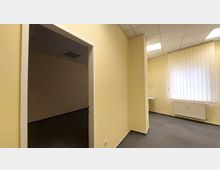 Leerstehender Innenraum eines Gebäudes mit cremefarbenen Wänden und einem grauen Teppichboden. Links führt eine Türöffnung in einen weiteren dunkleren Raum, rechts befindet sich ein großes Fenster mit vertikalen Lamellen und eine weiße Heizung darunter.