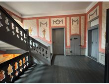 Treppenhaus in einem historischen Gebäude mit bemalten Wänden in Rot- und Beigetönen, dekorativen Ornamenten und grau gestrichenen Türen. Das Geländer der Holztreppe zeigt ein Muster mit abgerundeten Aussparungen, und der Boden ist aus dunklem Holz.