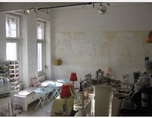 Heller Innenraum eines kleinen Cafés mit Vintage-Dekor. Zu sehen sind weiße Möbel, kleine runde Tische mit Blumenvasen, rote Lampenschirme mit Tupfen und eine Theke mit Geschirr und Snacks. Im Hintergrund sind große Fenster und eine helle Wand mit leicht erkennbaren Wandmalereien.