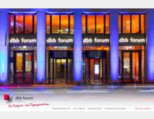 Frontansicht des dbb forum Berlin, eines modernen Kongress- und Tagungszentrums mit großer Glasfassade und leuchtender farblicher Beleuchtung in Orange- und Blautönen. Der Eingangsbereich verfügt über automatische Drehtüren und ist von beleuchteten Säulen eingerahmt.