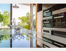 Moderne Küche mit einer Kücheninsel aus glänzendem Material, auf der eine dekorative Glasschale steht. Im Hintergrund große Fenster mit Blick ins Freie, umgeben von grünen Pflanzen, und integrierte Küchengeräte auf der rechten Seite.