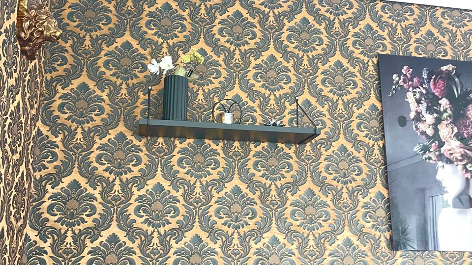 Wohnzimmer mit einer gold-schwarz gemusterten Tapete im Barockstil, einem grauen Ecksofa mit Kissen, einem Wandregal mit Dekoration wie einer Vase und kleinen Objekten sowie einem großen gerahmten Blumenbild an der Wand.