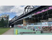 Eine urbane Szenerie mit einer Metallbrücke, die von Gerüsten und Wänden mit Graffiti flankiert wird. Im Vordergrund befinden sich ein grüner Spielbereich und zwei Personen, die miteinander sprechen, sowie ein Wohngebäude und Bäume im Hintergrund.
