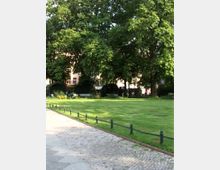 Grüne Parkanlage mit gepflegtem Rasen, umgeben von Bäumen und Büschen. Im Hintergrund sind Sitzbänke und teilweise Wohngebäude sichtbar, ein gepflasterter Weg verläuft am linken Bildrand.