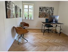 Helles, modernes Büro mit Fischgrätparkett, einem schwarzen Schreibtisch mit Computer, einem ergonomischen Stuhl und zwei braunen Sesseln an der Wand. Kunstwerke an den weißen Wänden und ein Fenster mit Blick auf grüne Vegetation sind sichtbar.
