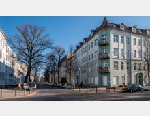 Straßenszene in einem Wohngebiet mit mehrstöckigen Gebäuden im klassischen Stil, teilweise mit grüner Fassade und Balkonen. Die Straße ist von kahlen Bäumen gesäumt, parkende Autos sind entlang des Gehwegs zu sehen, und der Himmel ist klar und blau.