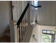 Eine einfache, helle Innentreppe mit Holzstufen, weißen Geländern und einem dunklen Handlauf. Im Hintergrund sind eine weiße Tür, ein Fenster sowie ein offener Eingang mit Blick auf einen gepflasterten Außenbereich sichtbar.