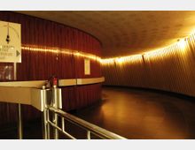 Ein geschwungener, beleuchteter Innenraum mit einer Wand aus poliertem Holz und vertikalen Leisten an der gegenüberliegenden Seite. Im Vordergrund sind eine Edelstahlabsperrung und ein kleines Schild mit einem Hinweis zum Panoramacafé und Aufzug sichtbar.