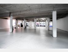 Großzügiger Innenraum eines leerstehenden Gebäudes mit Betonstützen, grauen Fliesenboden und weiß gestrichenen Wänden. Große Fensterfronten lassen Tageslicht herein und bieten Ausblick auf eine Straße mit geparkten Autos.