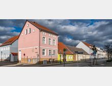 Straßenszene mit Reihenhäusern in Pastellfarben, darunter rosa, blau und gelb, mit roten Ziegeldächern und weißen Fensterrahmen, unter einem bewölkten Himmel. Im Vordergrund befinden sich Straßenschilder, eine Bushaltestelle und ein Baum.