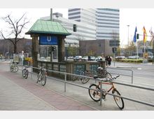 U-Bahnhof-Eingang "Theodor-Heuss-Platz" mit grünem Dach in einer städtischen Umgebung; im Vordergrund stehen Fahrräder an Geländern, im Hintergrund sind moderne Gebäude, Autos und Straßen mit Ampeln sichtbar.