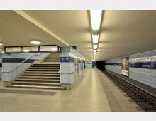 U-Bahn-Station mit blauen Wandfliesen, einer Treppe zum Ausgang, einem Bahnsteig mit Beleuchtung und Gleisen auf der rechten Seite. An der Wand befinden sich Werbeplakate und der Stationsname „Frankfurter Tor“.