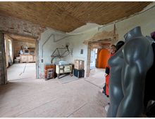 Ein unfertiger Innenraum mit sichtbaren Ziegelwänden, einem Boden aus einfachen Holzplatten und einer Decke mit Schilfrohrelementen. Der Raum enthält alte Möbel, Kartons, Dekorationsobjekte und mehrere Schaufensterpuppen; Türen führen in angrenzende Räume mit ähnlichem Zustand.