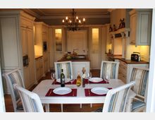 Klassische Küche mit cremefarbenen Einbauschränken, einer zentralen Kücheninsel und einer hängenden Metall-Kronleuchterlampe. Im Vordergrund steht ein gedeckter Esstisch mit vier Stühlen, weißen Tellern, roten Platzsets und Flaschen Wein sowie Öl.