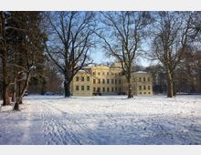 Gelbes, mehrstöckiges Herrenhaus im neoklassizistischen Stil, umgeben von schneebedecktem Park mit kahlen, großen Bäumen unter klarem, blauem Himmel. Im Vordergrund sind Fußspuren im Schnee sichtbar.