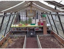 Innenansicht eines kleinen Gewächshauses mit geneigten Glaswänden und einem Tisch im Hintergrund. Auf dem Tisch stehen Körbe, Töpfe und Gartenwerkzeuge, während unten Beetanlagen mit Erde und Rankhilfen sichtbar sind.