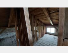 Schmaler Dachbodenraum mit Holzbalken und Holzwänden, dazu eine schräge Decke mit sichtbarer Holzverschalung. Am Ende des Raumes befindet sich ein kleines Fenster, das Tageslicht hereinlässt.