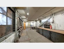 Industrieartiger Raum mit grober Einrichtung: Holzboden, Metallarbeitsflächen und Regalen an den Wänden. Eine vergitterte Fensterfront sowie freiliegende Rohre und Leitungen verstärken den funktionalen Charakter des Raumes; ein Feuerlöscher steht in einer Ecke.