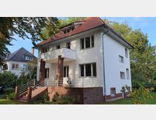 Zweigeschossiges Einfamilienhaus mit weiß verputzter Fassade und rotem Satteldach, einer erhöhten Veranda mit Klinkersteinsockel und Metallgeländer, umgeben von Bäumen und einem Garten. Im Hintergrund ist ein weiteres Wohnhaus mit Dachgauben zu sehen.