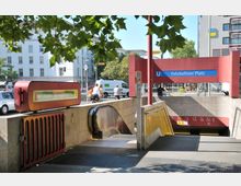 Eingang zur U-Bahn-Station „Fehrbelliner Platz“ in Berlin, mit einer Rolltreppe und Treppenstufen, umgeben von roten und beigen Steinwänden. Im Hintergrund sind ein Baum, parkende Autos und umliegende Gebäude zu sehen.