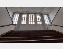 Holztreppe mit dunklen Stufen führt nach oben zu einer hohen Wand mit fünf großen, rechteckigen Fenstern, die Tageslicht durchlassen. Die Wände und die Fensterrahmen sind weiß, und die Decke zeigt einfache Zierleisten.