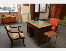 Ein vintage eingerichtetes Büro mit einem alten Holztisch mit grünem Ledereinsatz, darauf ein schwarzes Wählscheibentelefon. Im Raum stehen mehrere gepolsterte Stühle, ein kleiner Beistelltisch und ein Sideboard vor einer leicht beschädigten Wand mit großem Fenster.