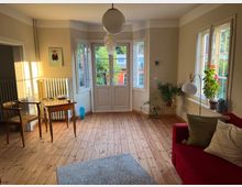 Helles Wohnzimmer mit Holzdielenboden, einer roten Couch, einem kleinen Holzschreibtisch und mehreren Zimmerpflanzen. Große Fenster und eine Glastür lassen viel Tageslicht herein und bieten Blick ins Grüne.