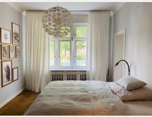 Helles Schlafzimmer mit einem großen Fenster, weißen Vorhängen und Blick auf eine grüne Außenanlage. An der linken Wand hängen eingerahmte Bilder, und im Vordergrund steht ein Bett mit heller Bettwäsche, neben dem sich eine Leselampe befindet.