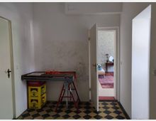 Ein schlichter Flur mit hellen Wänden und einem Boden aus schwarz-gelb karierten Fliesen. Links steht ein kleiner Tisch mit gelben Getränkekisten und einer roten Kabelrolle, rechts führt eine geöffnete Tür in einen angrenzenden Raum mit gemusterten Tapeten, einem roten Teppich und einem kleinen Tisch mit Spiegel.