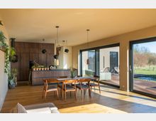 Moderne Wohnküche mit Holzboden, einer Kücheninsel aus dunklem Holz, hängenden Lampen und einem Esstisch mit Stühlen aus Holz. Große Glasfronten bieten Ausblick auf eine Terrasse und einen Gartenbereich.