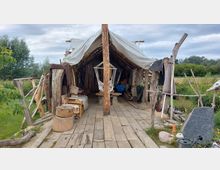 Ein rustikaler Unterstand aus Holz mit offener Front, einer Überdachung aus Plane und einfachen Holzmöbeln. Der Boden besteht aus Holzdielen, umgeben von einer ländlichen Wiesenlandschaft mit vereinzelten Bäumen im Hintergrund.