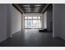 Ein großer, leerer Raum mit hohen Decken, Betonboden und sichtbaren Betonbalken. Vor einer großen Fensterfront, die Tageslicht hereinlässt, befinden sich eine graue Eckcouch und ein kleiner Holztisch.