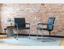 Zwei schwarze Stühle mit Metallgestell stehen auf einem hellgrauen Teppich vor einer roten Backsteinwand. Links daneben befindet sich ein kleiner Beistelltisch aus Holz und Beton mit einer Topfpflanze darauf.