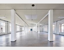 Großer, leerer Innenraum mit glattem Betonboden, weiß gestrichenen Wänden und mehreren quadratischen Säulen. Die rechteckigen Fenster entlang der Wand lassen viel Tageslicht herein, während die Decke mit eingelassenen Lichtfeldern und perforierten Paneelen gestaltet ist.