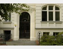 Eingangsbereich eines historischen Wohnhauses mit hellbeiger Fassade, verziertem Rundbogen über einer dunklen Holztür mit Glaseinsatz und der Hausnummer „20“. Vor dem Gebäude befinden sich ein kleiner Gartenbereich und ein schmiedeeiserner Zaun.