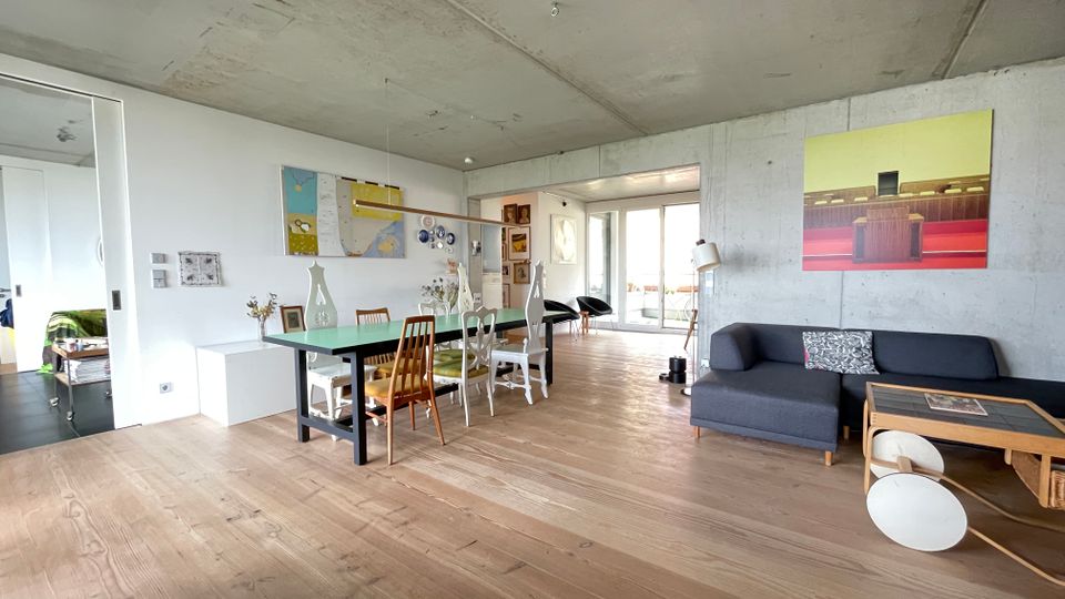 Wohnbereich in einem modernen Apartment mit offenem Grundriss, Betonwänden und hellem Holzboden. Eine graue Eckcouch, ein Esstisch mit unterschiedlich gestalteten Stühlen und große Fenster, die auf einen Balkon führen, bestimmen den Raum.