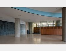 Ein moderner Innenraum mit glatten, hellen Bodenfliesen, einer Betonstütze und einer Wand aus Holzpaneelen mit Glaspartien darüber. Links ist eine graue Wand mit eingraviertem Text sichtbar, und die Decke hat eine geschwungene, blaue Einfassung.