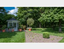Ein Garten mit einem kleinen weißen Pavillon, umgeben von Büschen und Blumentöpfen, daneben Metallstühle und ein Holztisch. Im Vordergrund ein frisch bepflanztes Beet, im Hintergrund Bäume und eine Holzbank.