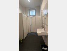 Öffentliche Waschräume mit weißen Trennwänden für Toilettenkabinen, einem Waschbecken an der Wand und einem kleinen, vergitterten Fenster oben in der Mitte. Der Boden ist mit dunklen Fliesen ausgestattet, und ein Mülleimer steht unter dem Waschbecken.