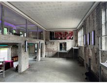 Industriehalle mit modernem Innenausbau: Ein zweistöckiger Raum mit unverputzten Ziegelwänden, großen Fenstern und Betonstützen. Es gibt offene Galerien mit Glasgeländern, eine Metalltreppe und violette Lichtakzente an den Wänden, die den Raum erhellen.