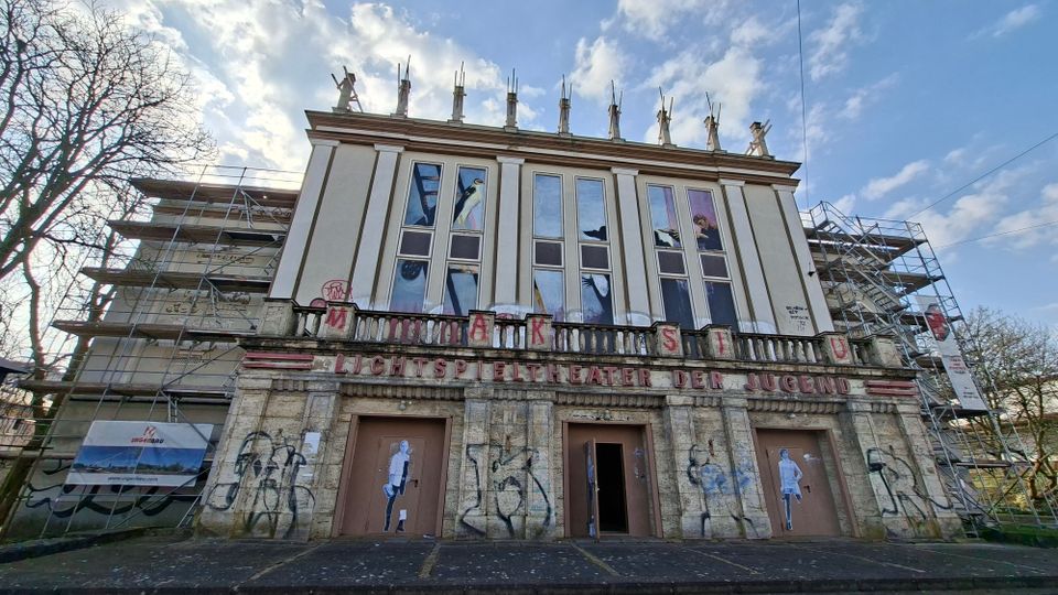 Frontansicht eines ehemaligen Lichtspieltheaters mit klassizistischer Fassade, hohen schmalen Fensterfeldern und Balustrade; über dem Eingang steht „LICHTSPIELTHEATER DER JUGEND“. Das Gebäude ist teilweise eingerüstet, die Eingänge sind verschlossen bzw. geöffnet, und die Außenwände zeigen Graffiti; davor verläuft eine breite, leere Vorfläche unter wolkigem Himmel.