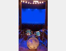Ein Schlagzeug in der Mitte einer Bühne steht im Vordergrund, während im Hintergrund ein leerer Zuschauerraum mit blauen Sitzen und blauer Beleuchtung zu sehen ist. Oben sind Bühnenscheinwerfer angebracht, die die Szene beleuchten.
