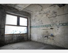 Verlassener, stark verwitterter Innenraum mit abblätterndem Putz, gewölbter Decke und freiliegenden Leitungen. An der gefliesten Wand mit blaugrünem Zierstreifen hängt ein altes Metallwaschbecken, daneben ein großes, milchig verschmutztes Fenster.