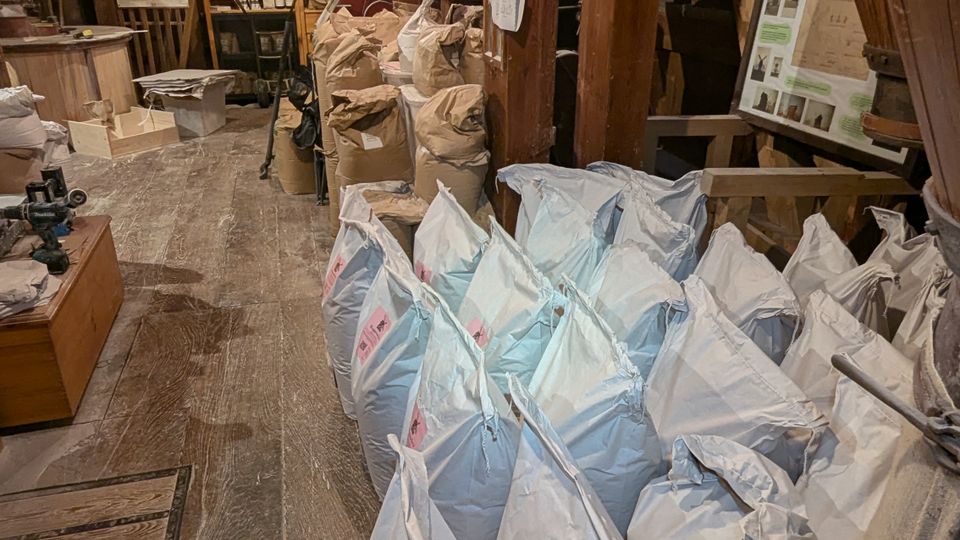 Innenansicht eines historischen Dachbodens oder Lagerraums mit freiliegenden Holzbalken, Holzfußboden und gedämpfter Beleuchtung. Entlang eines schmalen Gangs stehen zahlreiche große Säcke und Kisten, dazu hängen Infotafeln und Schilder wie in einer Ausstellung oder Werkstatt.