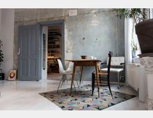 Ein minimalistischer Wohnraum mit einem Holztisch, umgeben von verschiedenen Stühlen, darunter ein weißer Kunststoffstuhl und ein Holzstuhl. Der Raum hat eine unverputzte Wand mit Patina, eine offene graue Tür, helle Holzböden und weiße Wände; im Hintergrund ein Regal mit Büchern und Kisten.